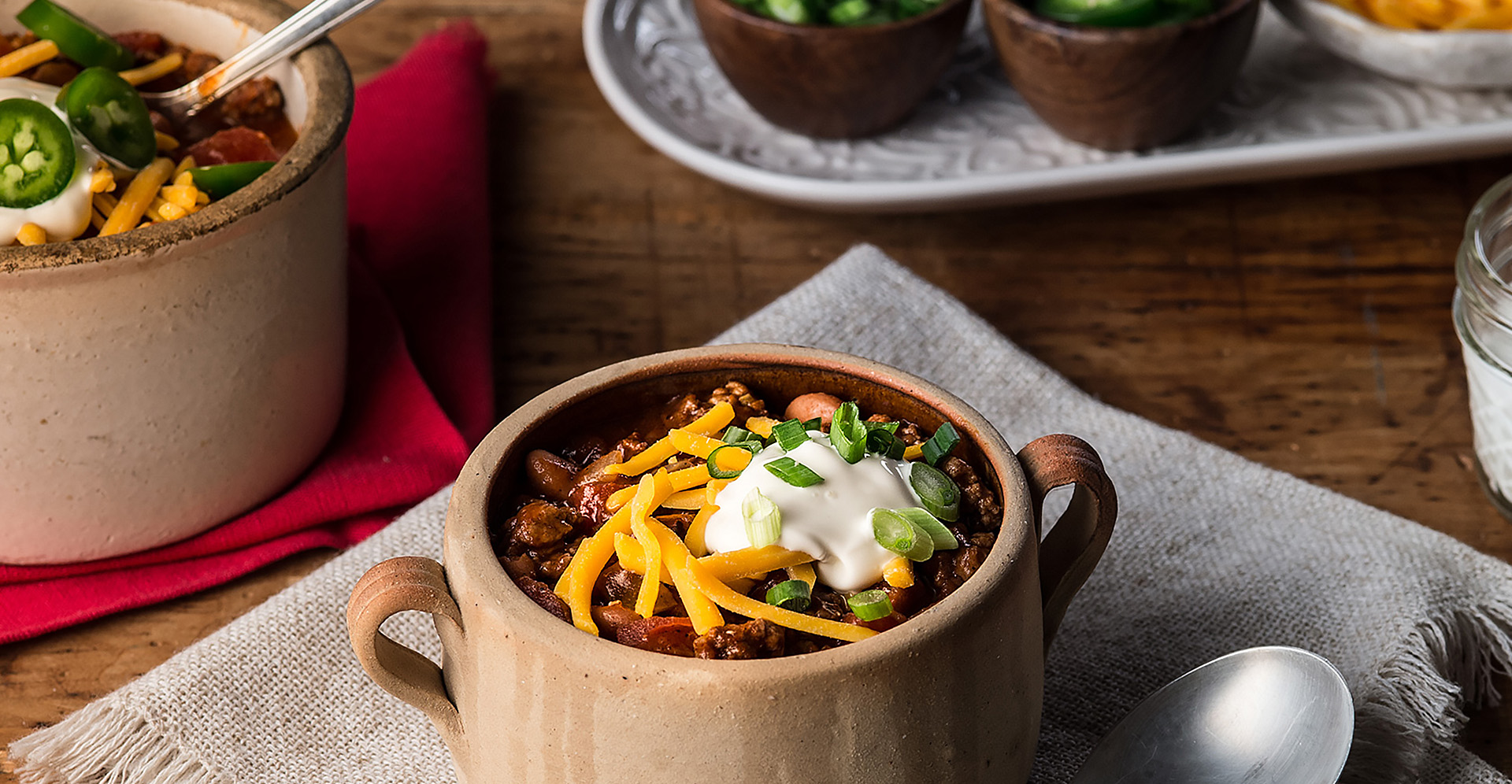 Bean and beef chili in small tan crock, garnished with shredded cheese, sour cream and green onions 