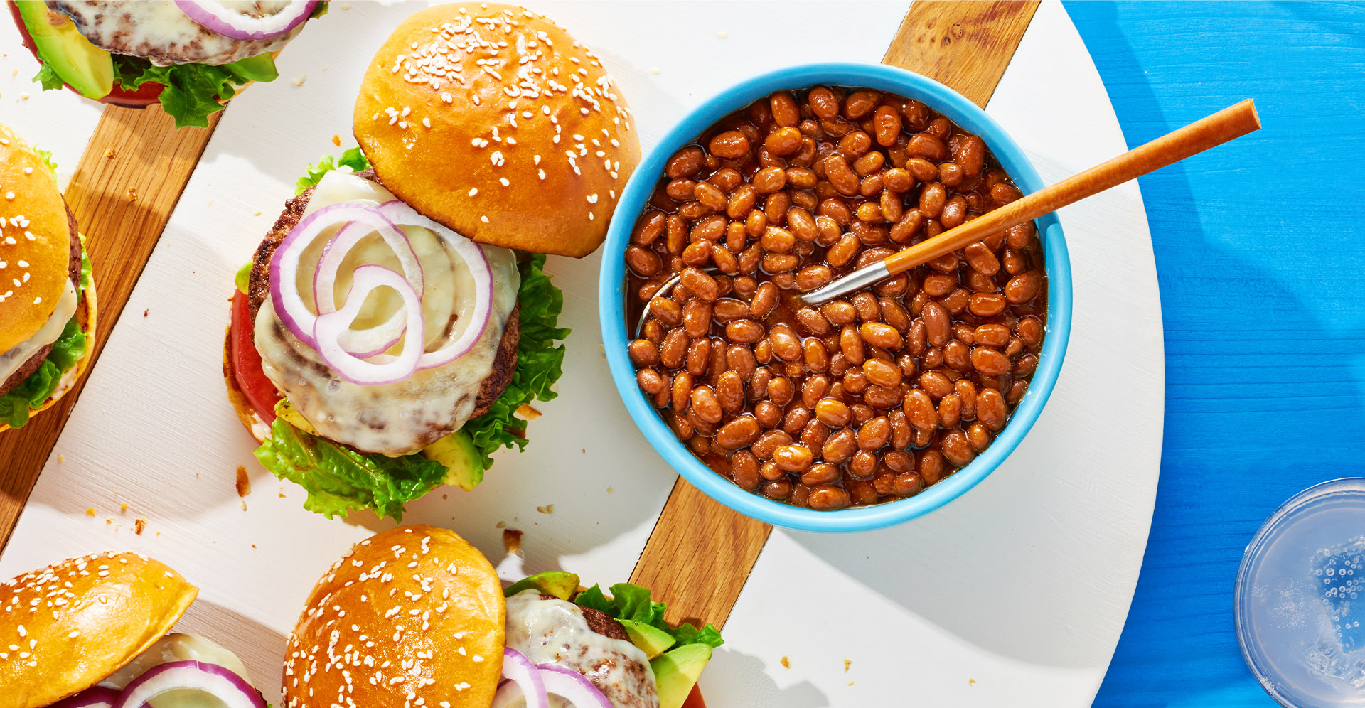  A blue bowl of Sweet Heat beans next to several burgers on a white plate.