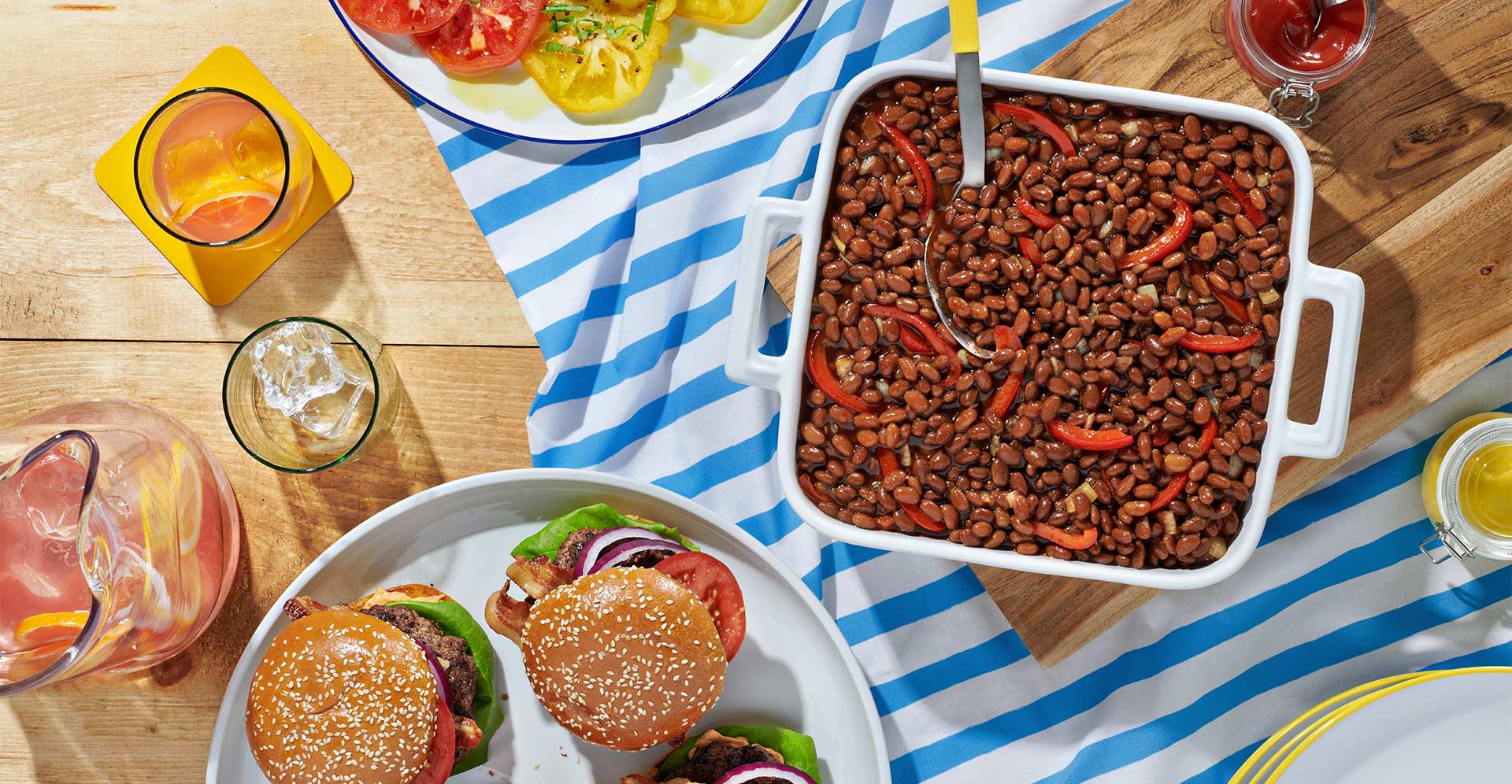 Top down tabletop view of a casserole dish with beans, onions and red peppers, along with a plate of burgers.