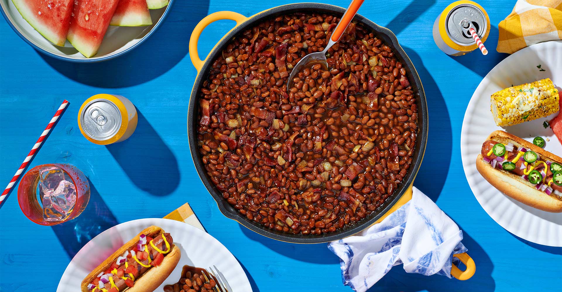 Top down view of baked beans (with bacon and onions) in an enamled pot surrouned by paper plates with hot dogs, pasta salad, corn on the cob elote, and watermelon. 